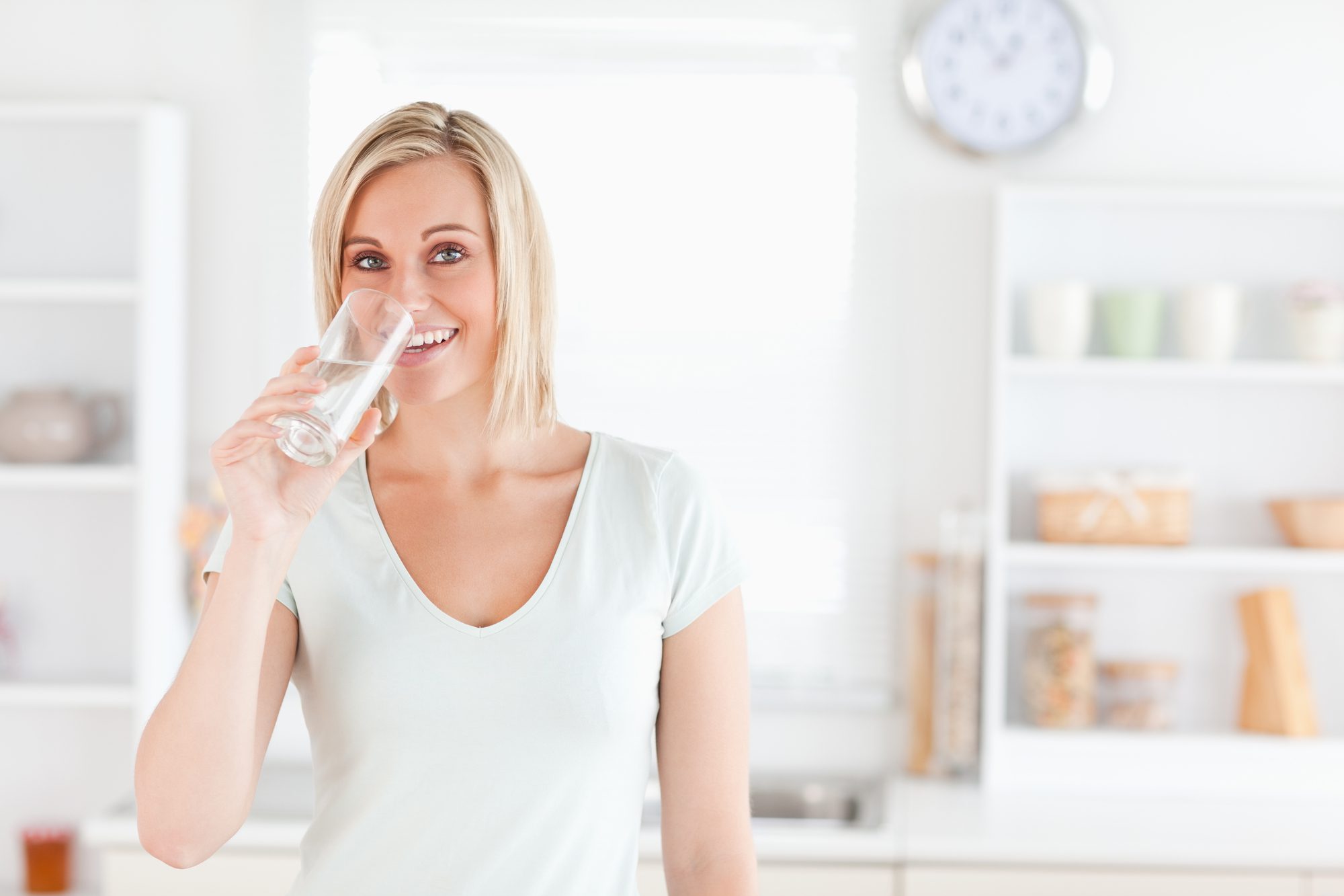 Charming woman drinking water while standing looks into camera in the kitchen